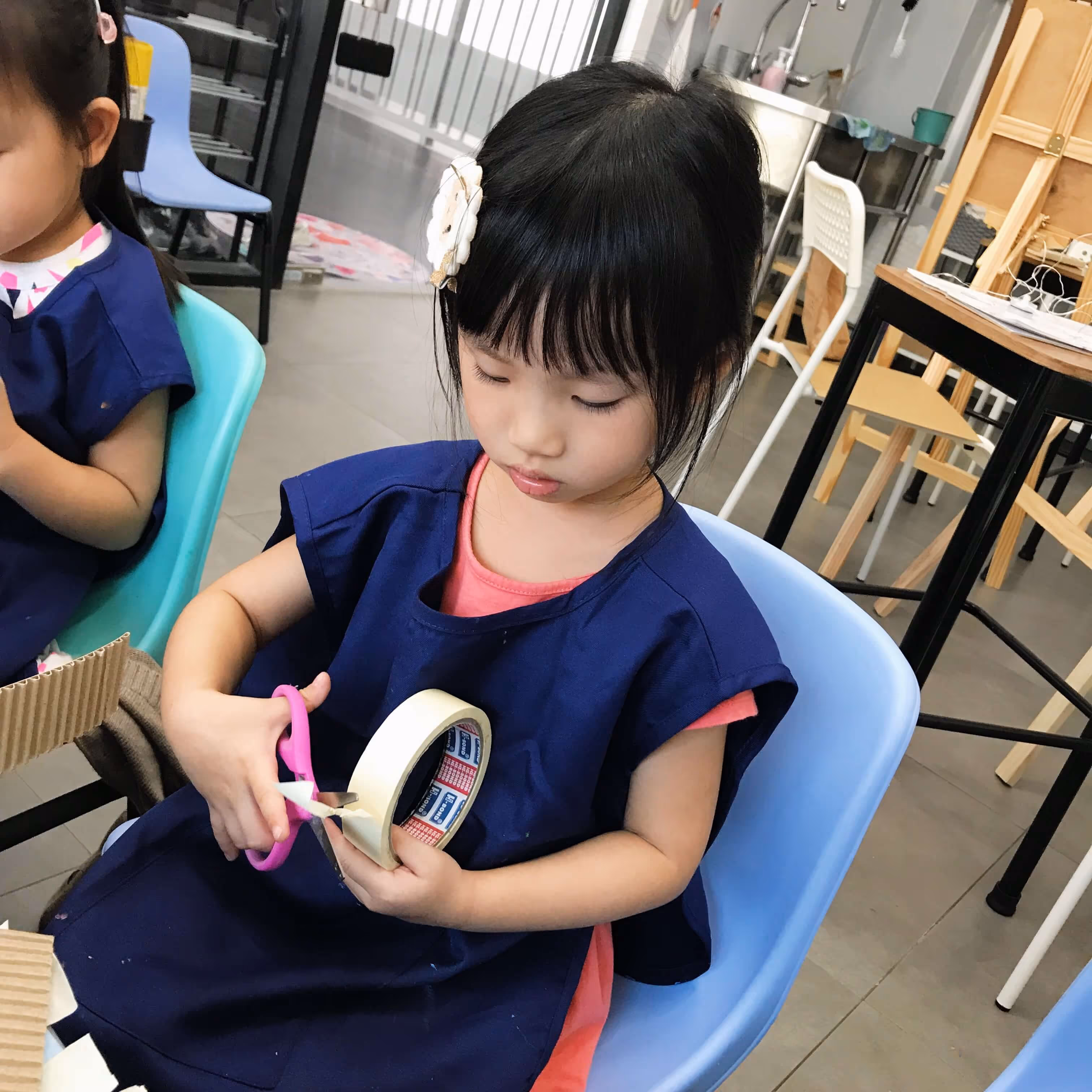 Young girl in a blue apron sitting on a light blue chair cutting masking tape with pink scissors in a classroom.