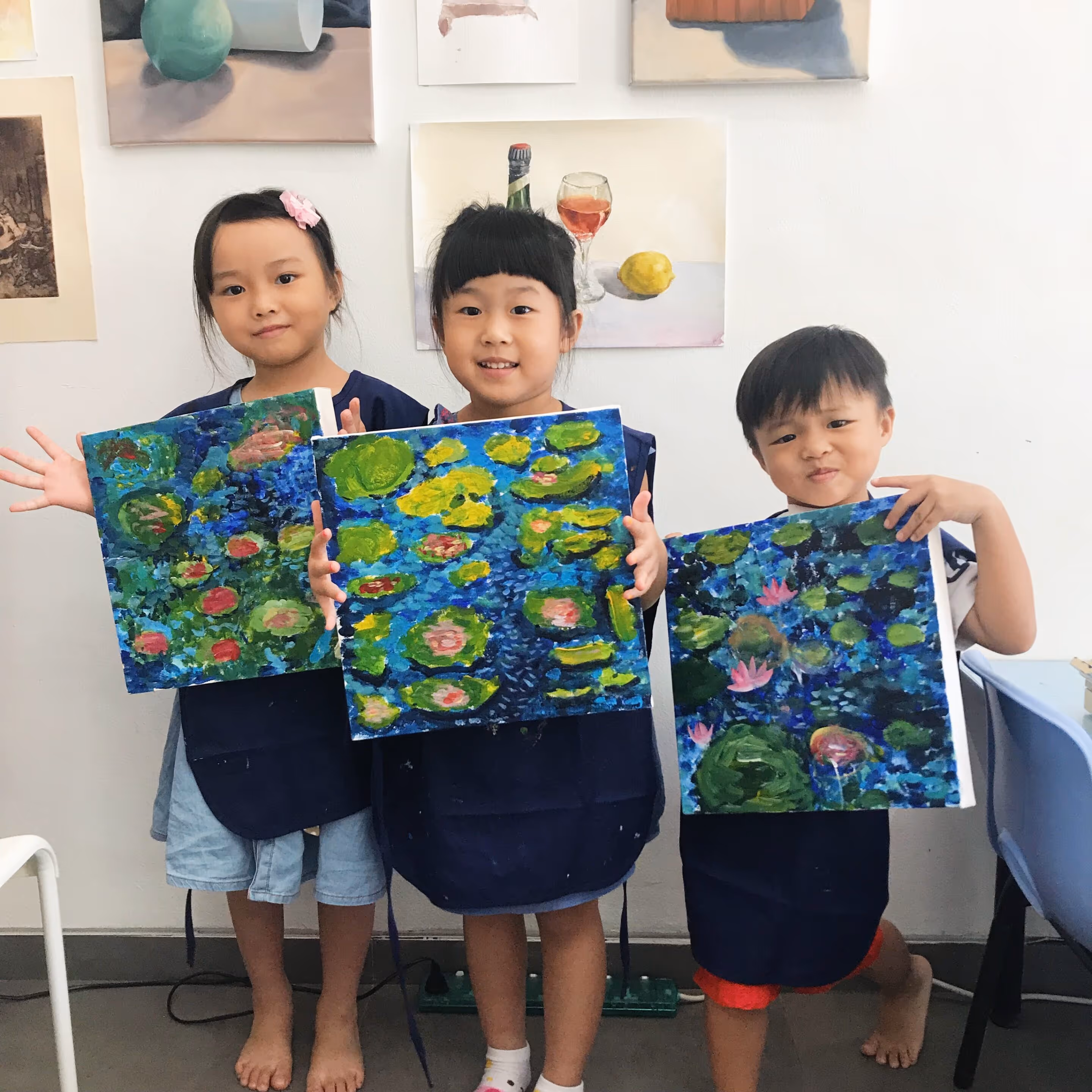 Three young children standing barefoot indoors holding colorful paintings of water lilies on blue backgrounds.