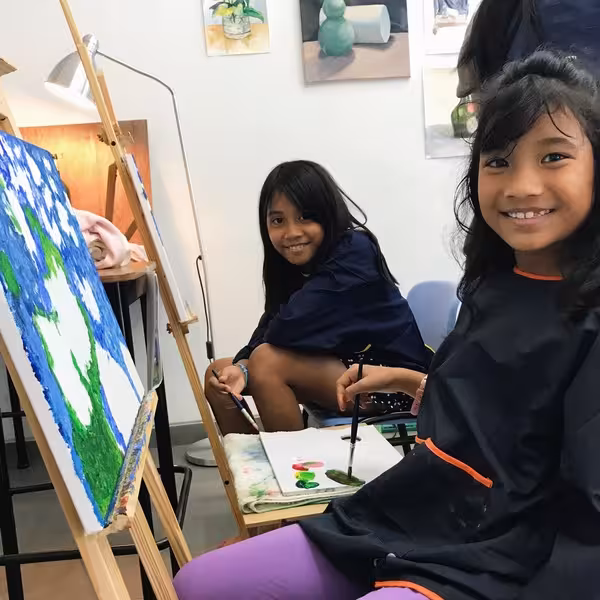 Two girls smiling while painting on canvases in an art class with colorful artwork and paintbrushes.