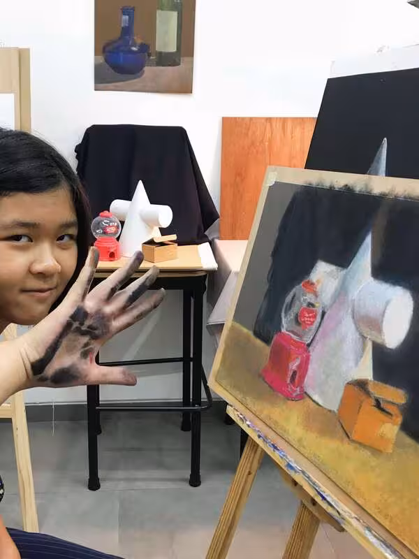 Young artist with charcoal-covered hands posing next to an easel displaying a still life pastel drawing of geometric shapes and bottles.