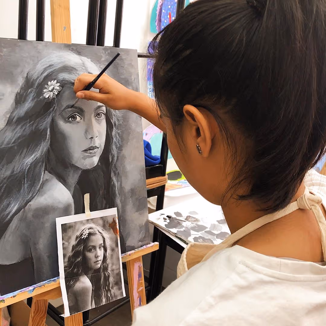 A female artist painting a grayscale portrait of a girl with long hair and a flower, using a reference photo taped to the easel.