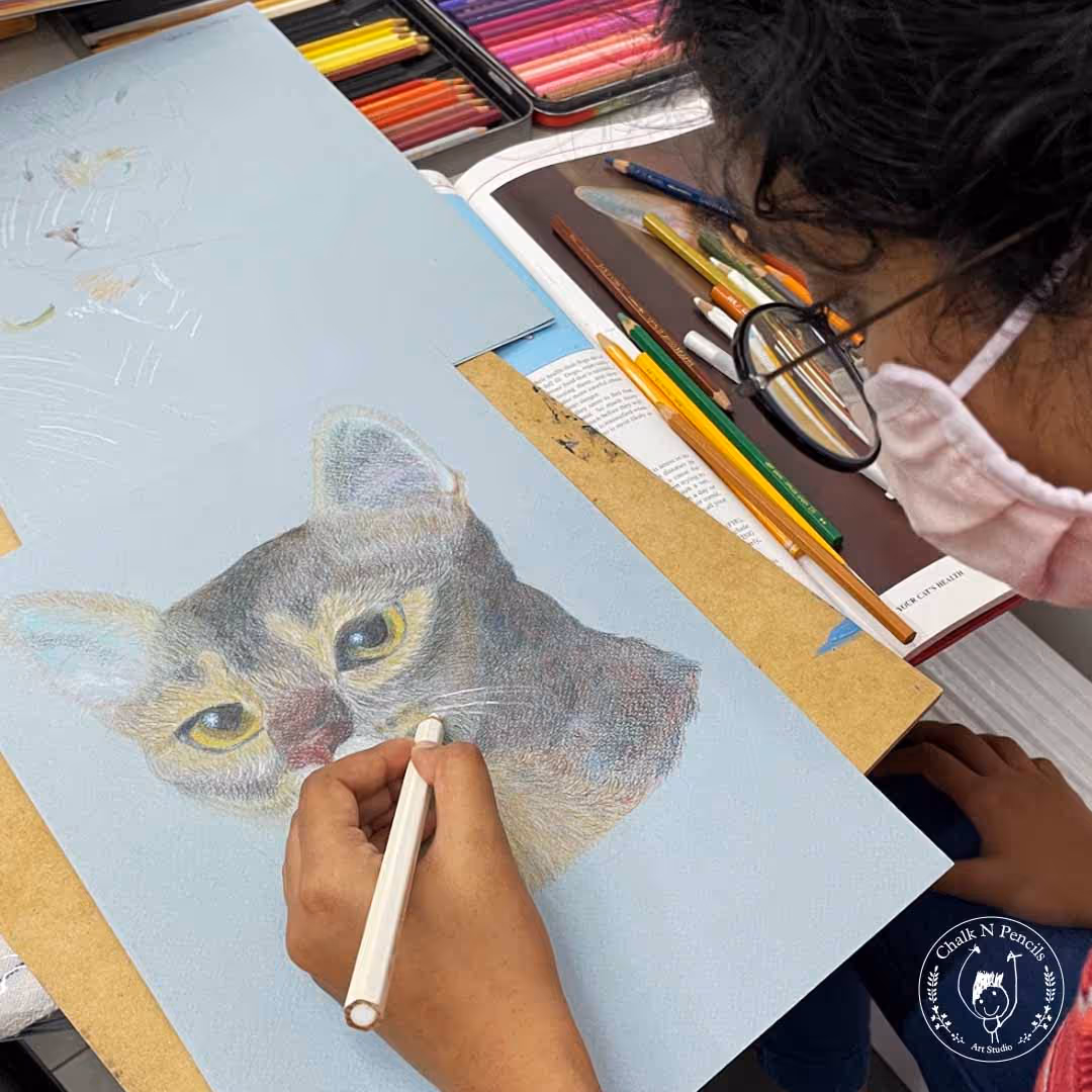 Person wearing glasses and a pink mask drawing a detailed colored pencil portrait of a cat on light blue paper.