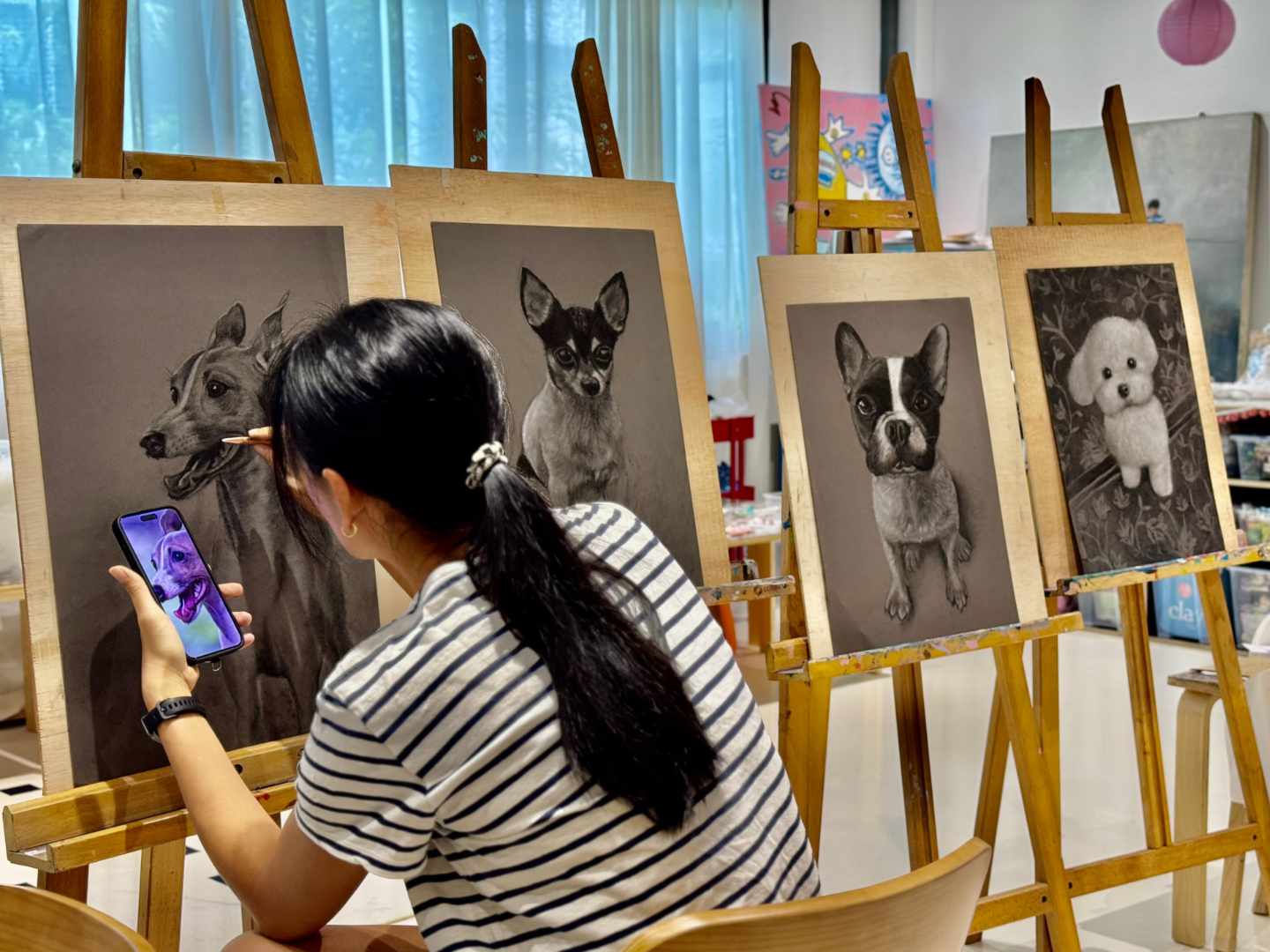 An artist takes a photo of their dog on a smartphone and turns it into a detailed charcoal drawing on an easel in a well-lit studio, alongside their other dog portraits.