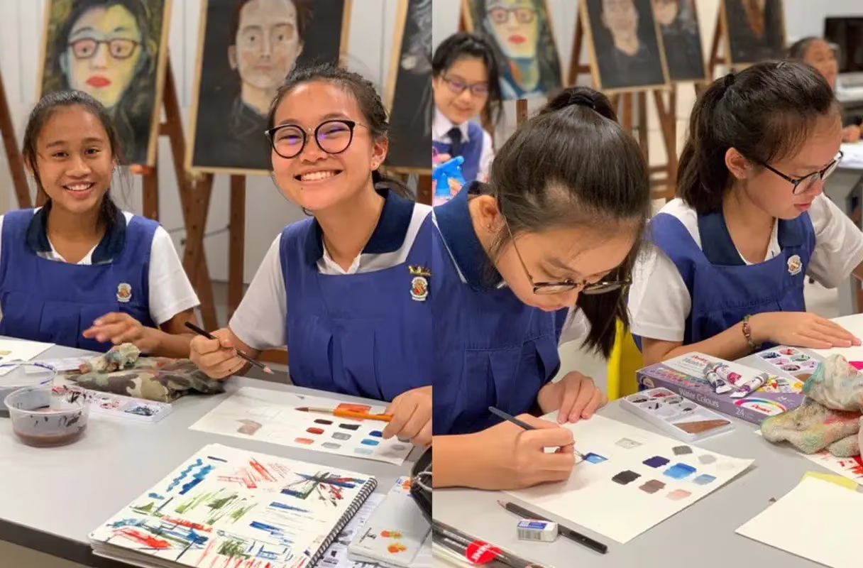 Students in blue uniforms painting and drawing with watercolors at a table in an art classroom with portrait artworks on easels in the background.