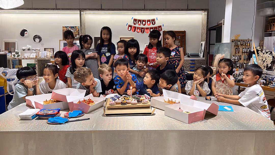 Group of children gathered around a table with cupcakes and party food celebrating a birthday with a Happy Birthday banner in the background.
