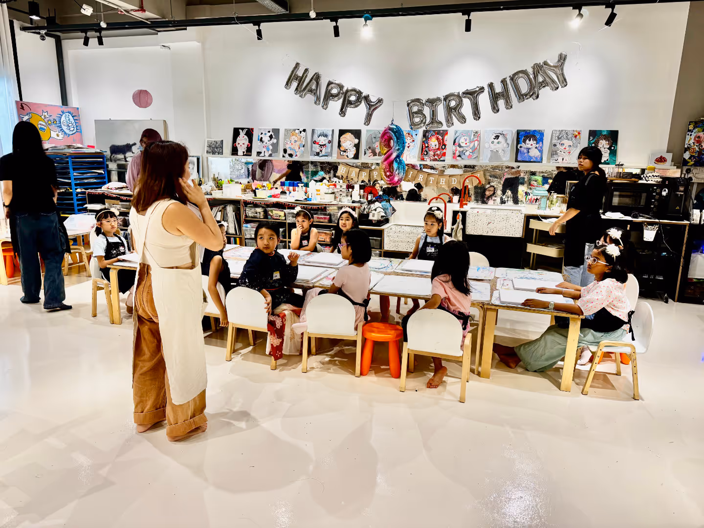 Children are seated around tables in a bright art studio decorated with 'Happy Birthday' balloons and artwork on the wall, with art teachers supervising.