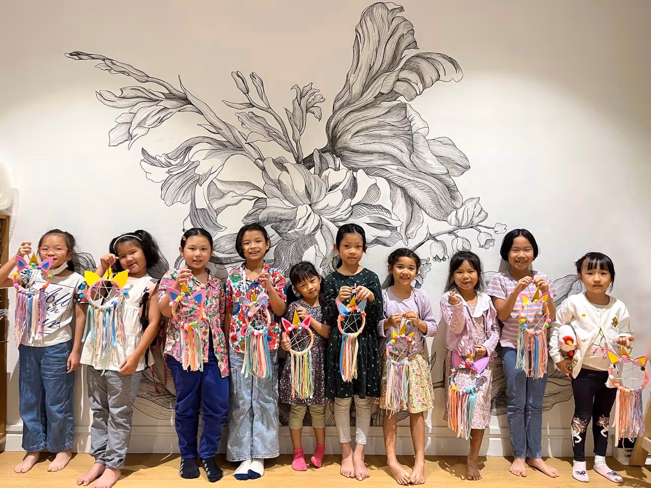 Ten children standing barefoot in front of a wall with floral mural, each holding colorful cat-themed dreamcatchers.
