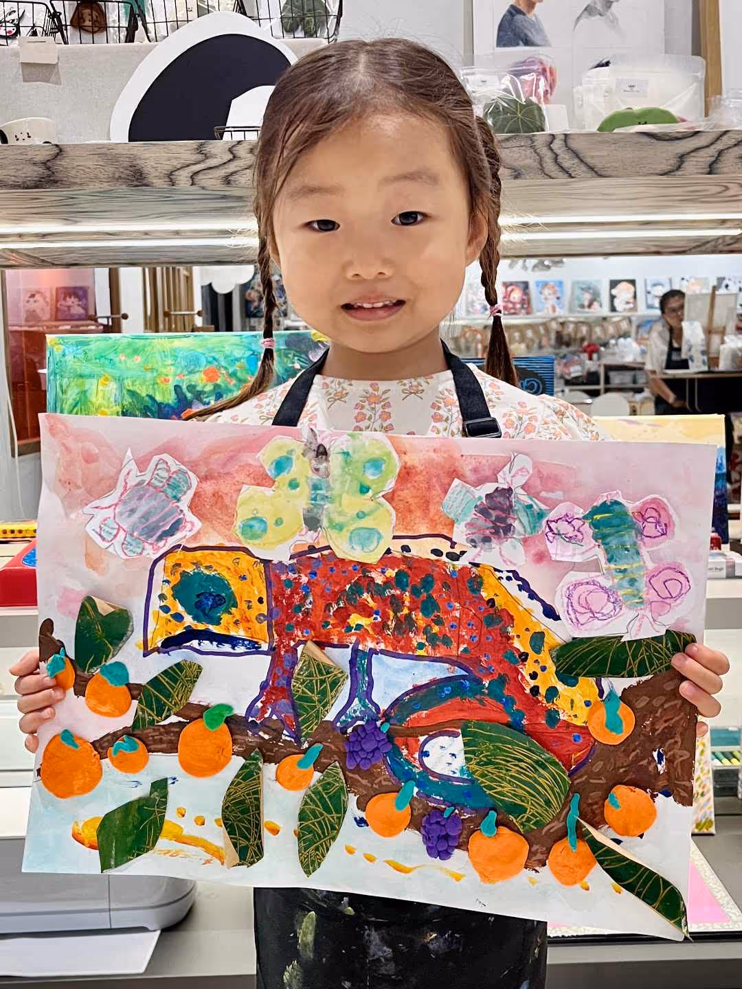 Young girl with braided hair holding a colourful painting of a chameleon on a branch with orange fruits and butterflies above.
