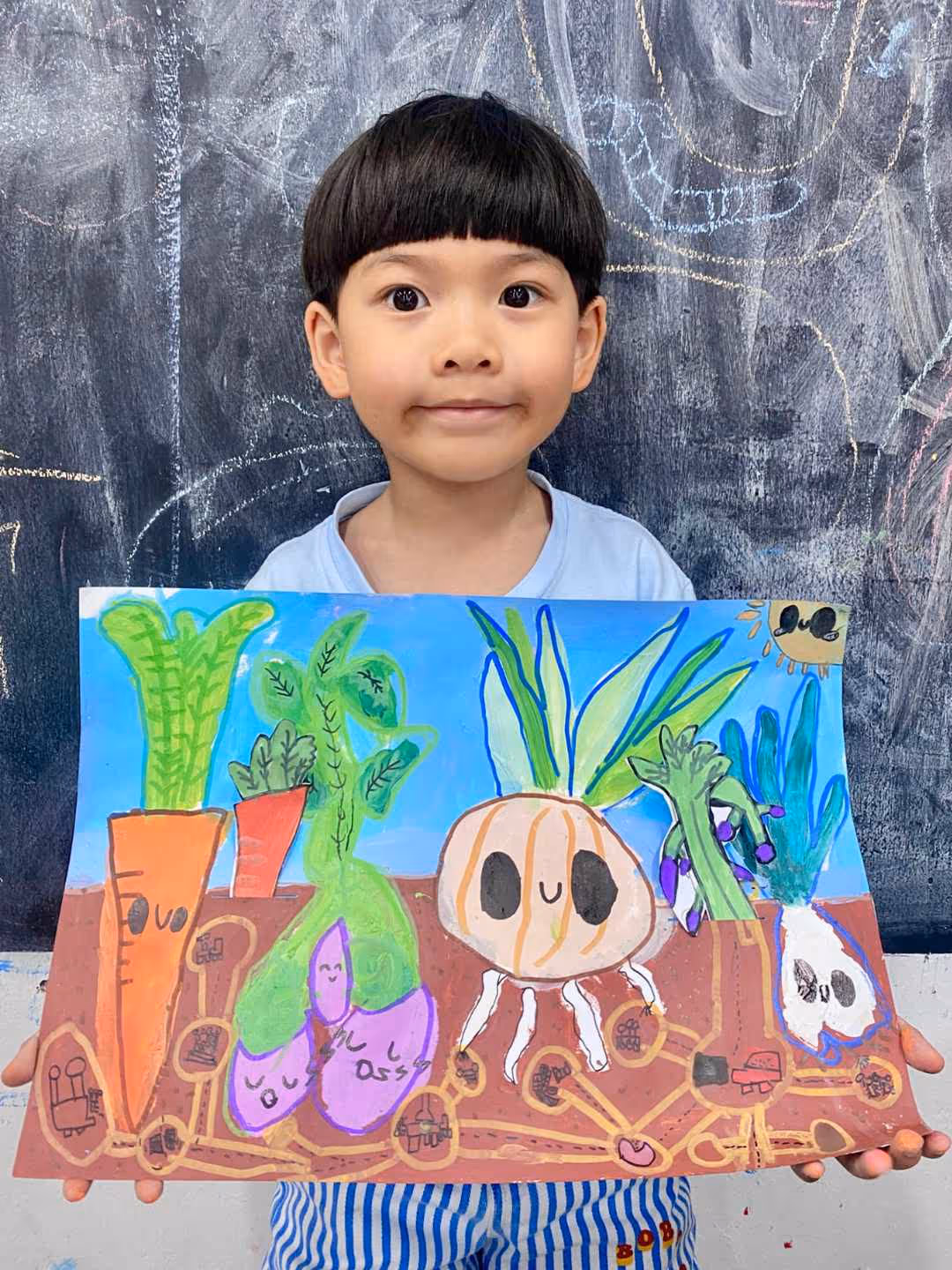 6-year-old boy holding a colourful drawing of root vegetables with smiling faces against a chalkboard background.