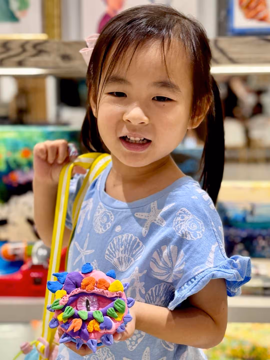 Young girl in a blue seashell-patterned shirt holding a colourful clay monster with one large eye.