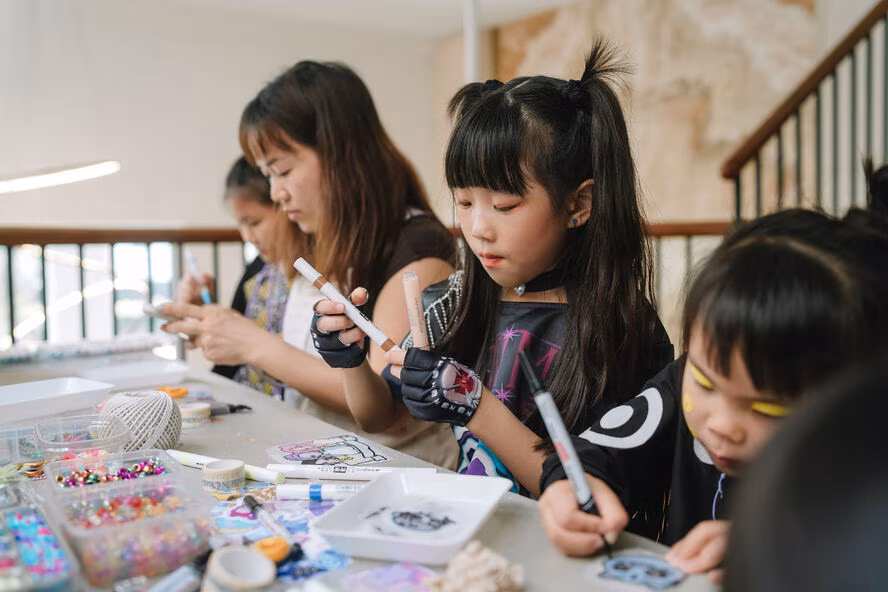 Children and an adult are focused on arts and crafts, using markers and decorative materials at a table.