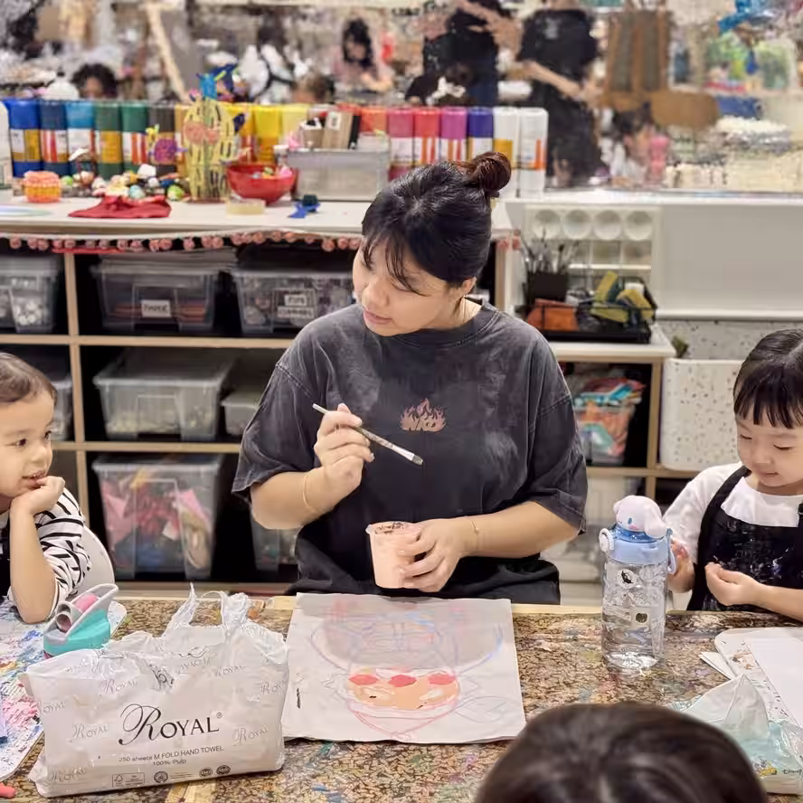 Ms Abby is holding a paintbrush and a cup of paint, sitting at a table with children engaged in an art activity at Chalk N Pencils Art Studio