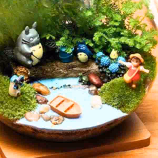 Miniature terrarium featuring Totoro figures, a small boat on a water-like surface, moss, and a girl figurine in a pink dress and hat.