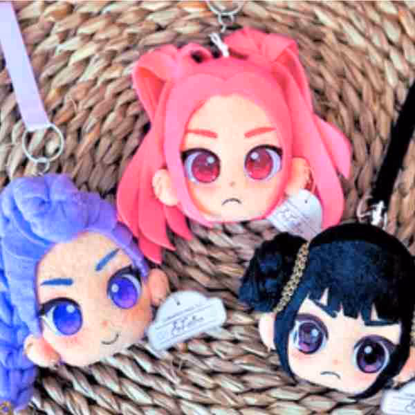 Three anime-style plush keychains with large expressive eyes placed on woven mat; one with pink hair, one with purple hair, and one with black hair.