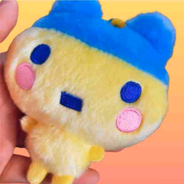 Hand holding a small Tamagotchi plush with a yellow body, blue hat with bear ears, blue eyes, and pink cheeks against an orange background.