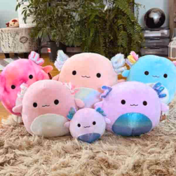 Six colorful, round plush axolotl toys with smiling faces in pastel pink, purple, peach, and blue shades arranged on a beige shaggy carpet indoors.