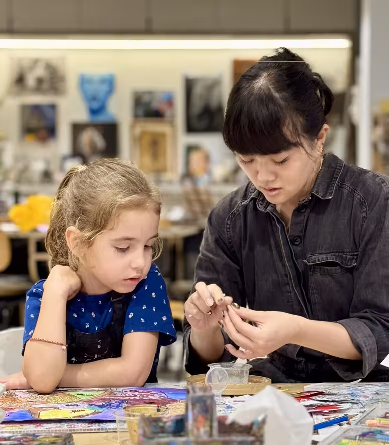 Ms Jessica instructs young girl on a project at a colourful table filled with art materials.