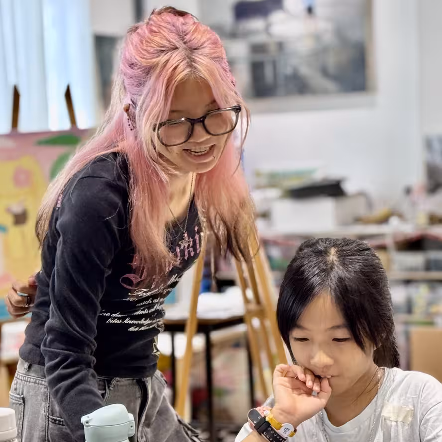 Ms Naomi with pink hair helping a young student in an art classroom with easels in background.