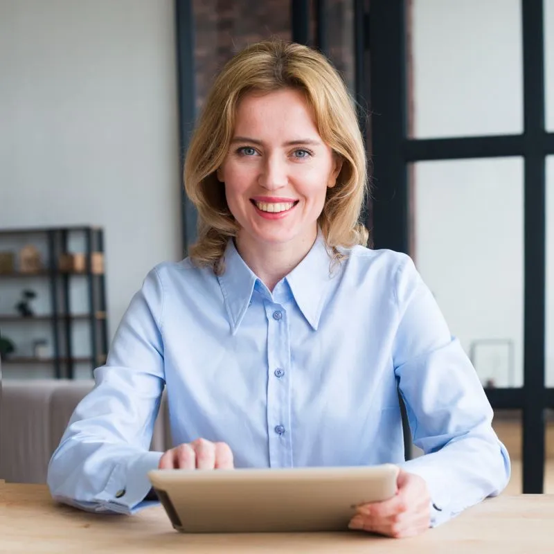 Smiling woman with blonde hair wearing a light blue shirt sitting at a table using a tablet.