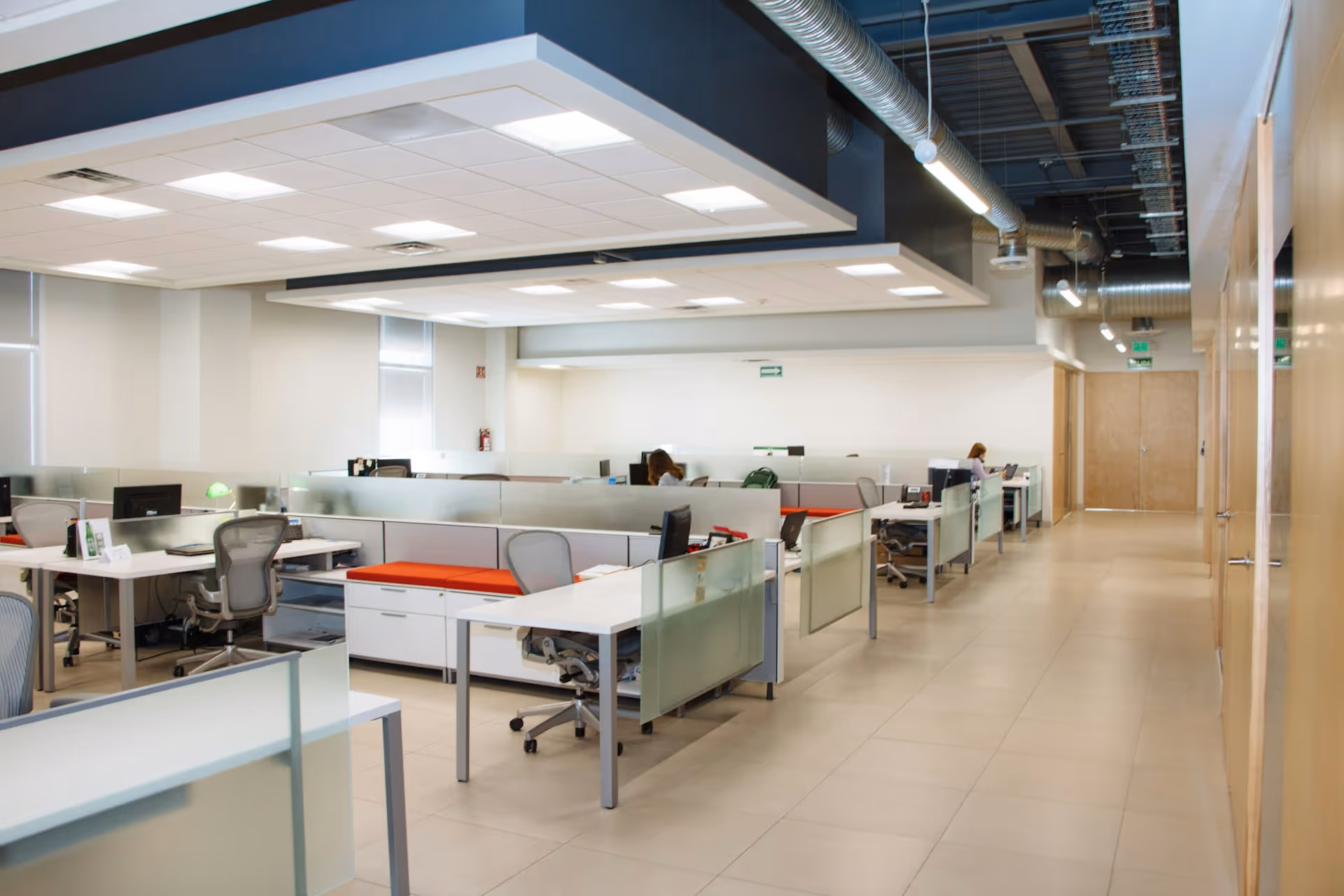 Modern office with individual workstations featuring white desks, ergonomic chairs, and frosted glass partitions under bright ceiling lights.