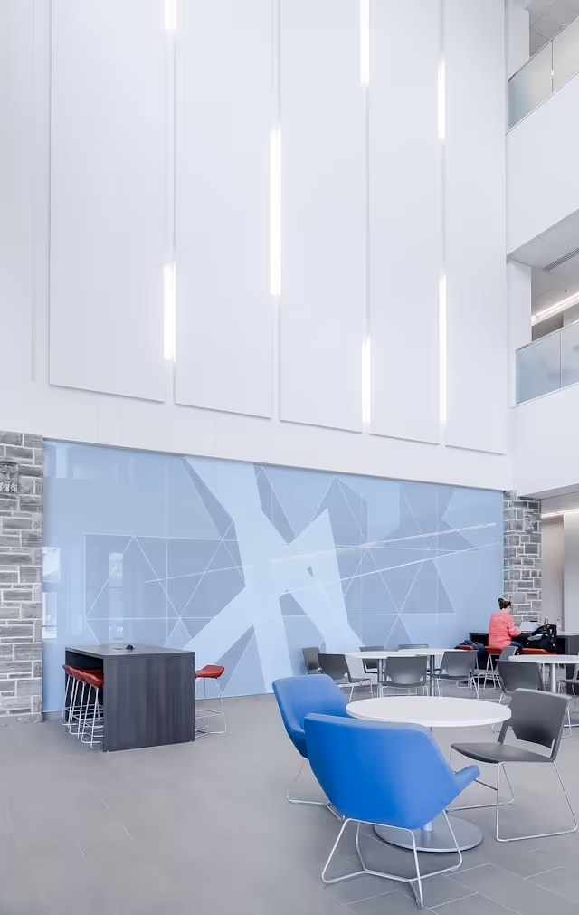 Modern open lounge area with blue lounge chairs, gray chairs, white round tables, and a large light-blue geometric wall.