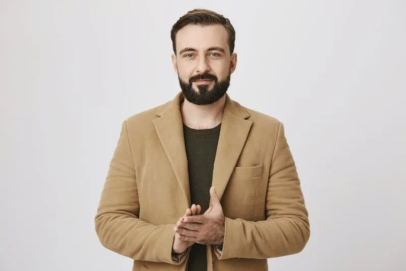 Bearded man in a brown blazer and dark shirt standing against a plain light background, clasping his hands.