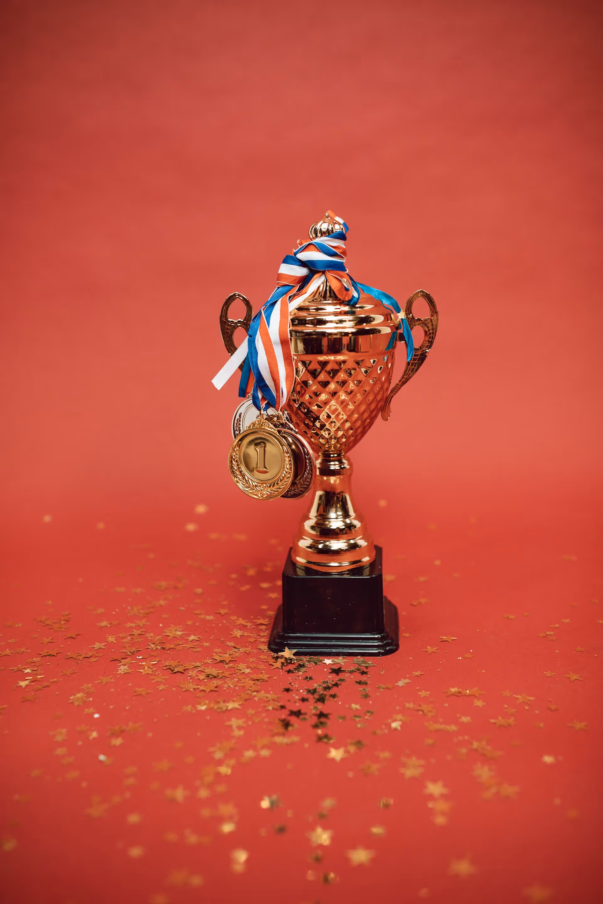 Gold trophy with multiple medals hanging from its top, placed on a red surface scattered with small gold star-shaped confetti.