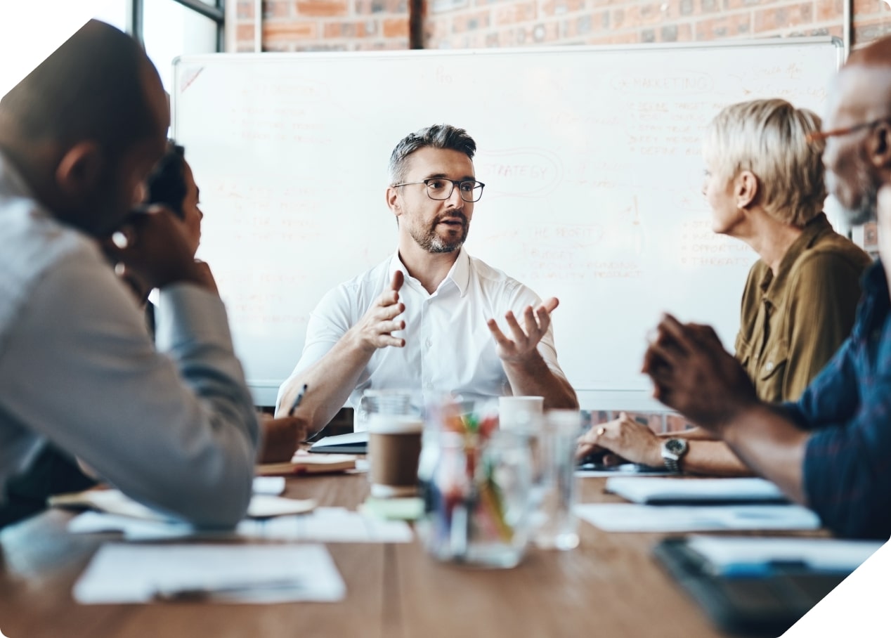 Man in glasses and white shirt speaking and gesturing during a team meeting with diverse colleagues around a table.