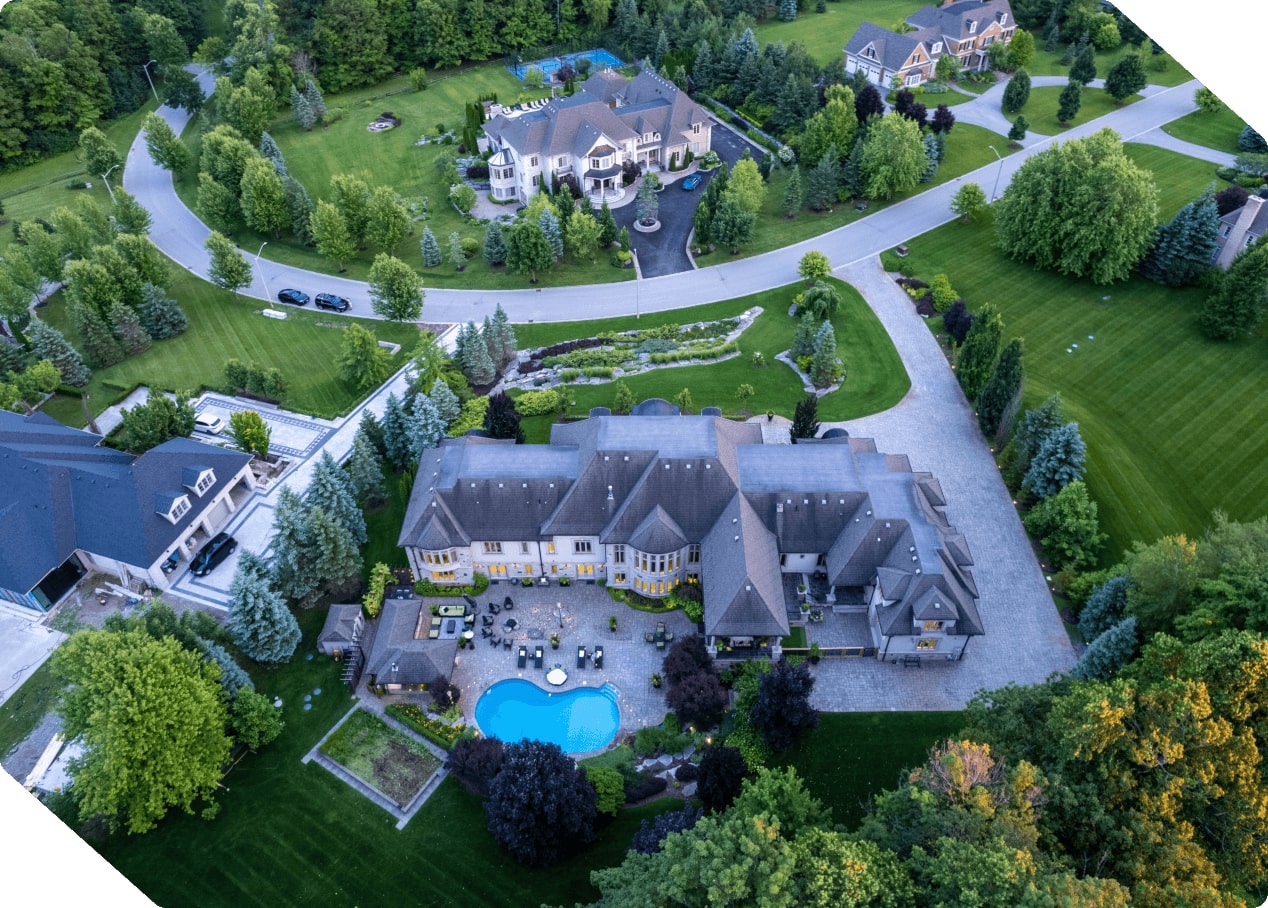 Aerial view of a large mansion with a backyard pool, surrounded by green lawns, trees, and other houses.