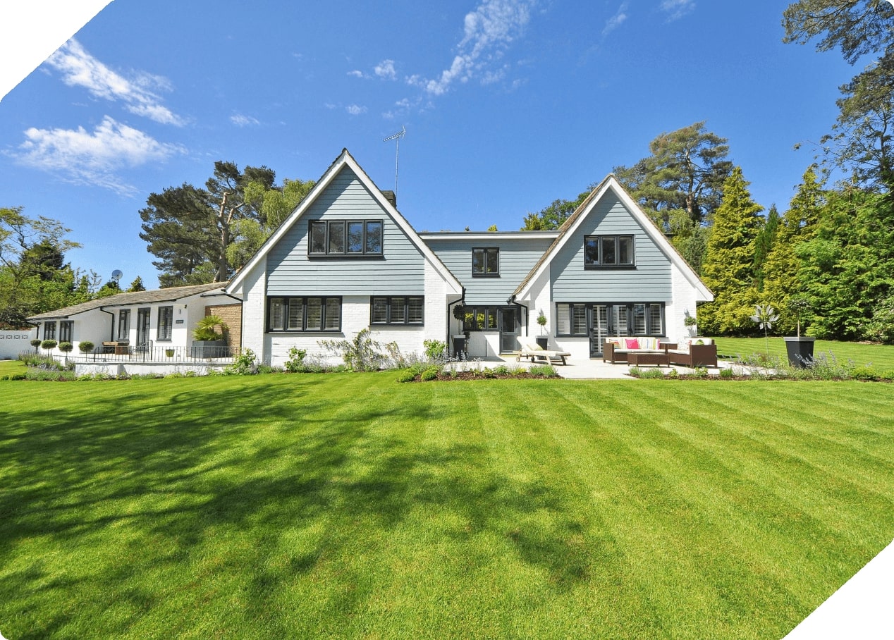 Large modern two-story house with blue and white exterior, surrounded by a lush green lawn and tall trees under a clear blue sky.