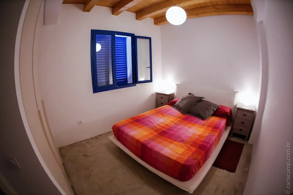 Dormitorio en Formentera con cama doble cubierta con sábana de cuadros naranjas, rojos y rosas, paredes blancas, ventana azul abierta, y techo con vigas de madera.