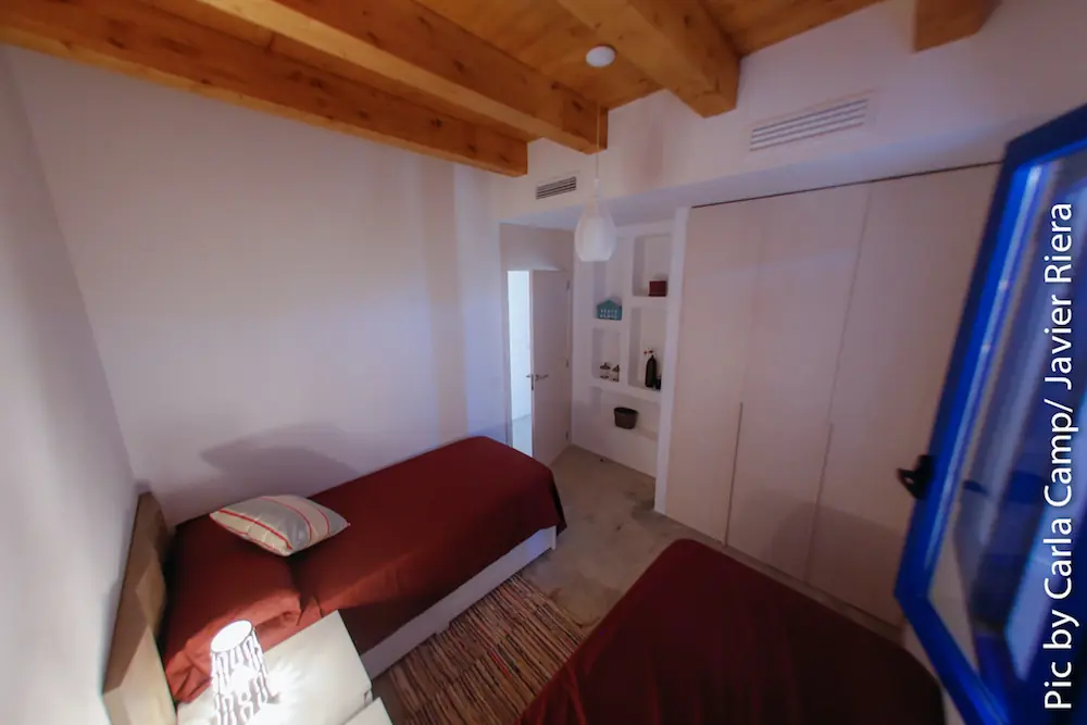 Dormitorio en Formentera con dos camas individuales, vigas de madera en el techo y ventana azul abierta.
