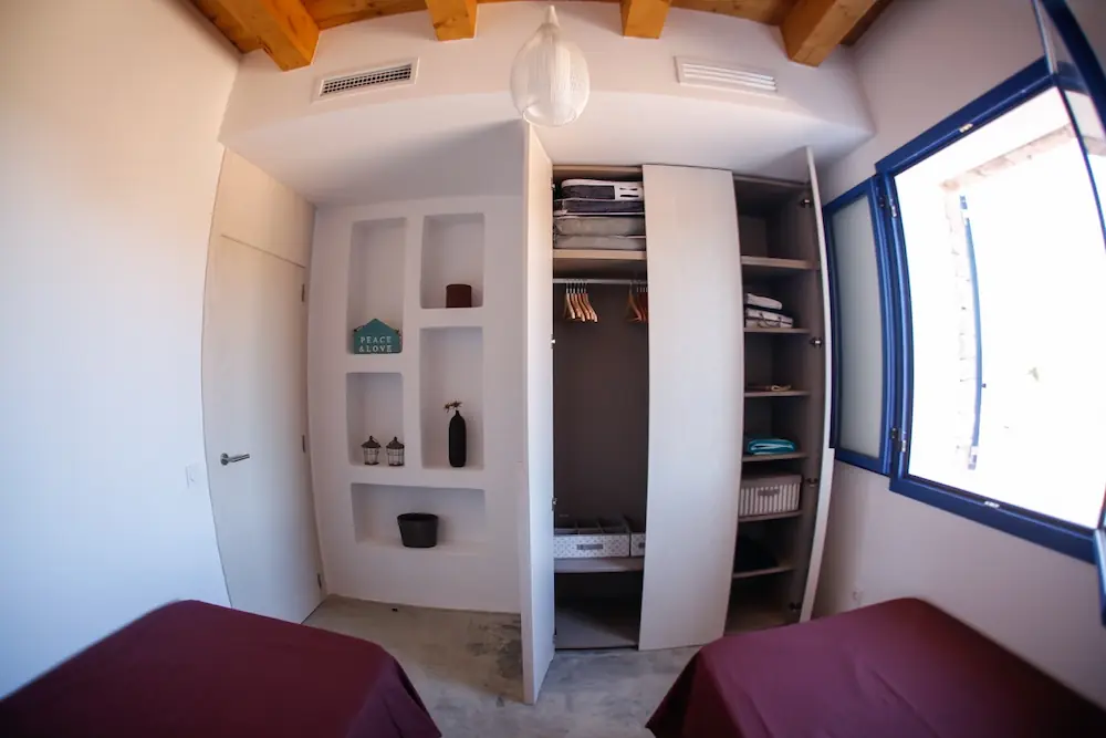 Habitación en El Pilar de la Mola con dos camas con mantas granate, armario abierto con estantes y perchas, estantería empotrada en la pared y ventana azul abierta.