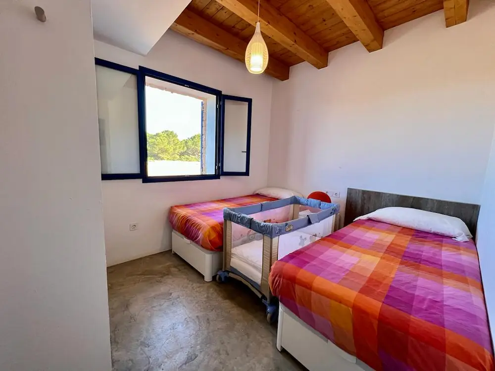 Dormitorio con dos camas individuales en Can Javi de Palma con ropa de cama a cuadros en tonos rojos, naranjas y rosas, y una cuna azul entre ellas, con luz natural y techo de madera.