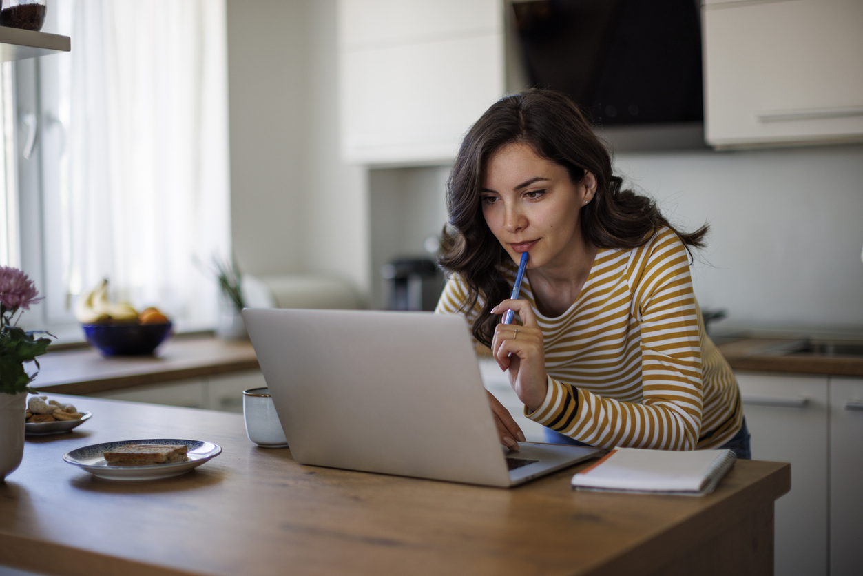 Young woman in a yellow striped shirt thoughtfully using a laptop at a kitchen island with a notebook and breakfast items nearby.