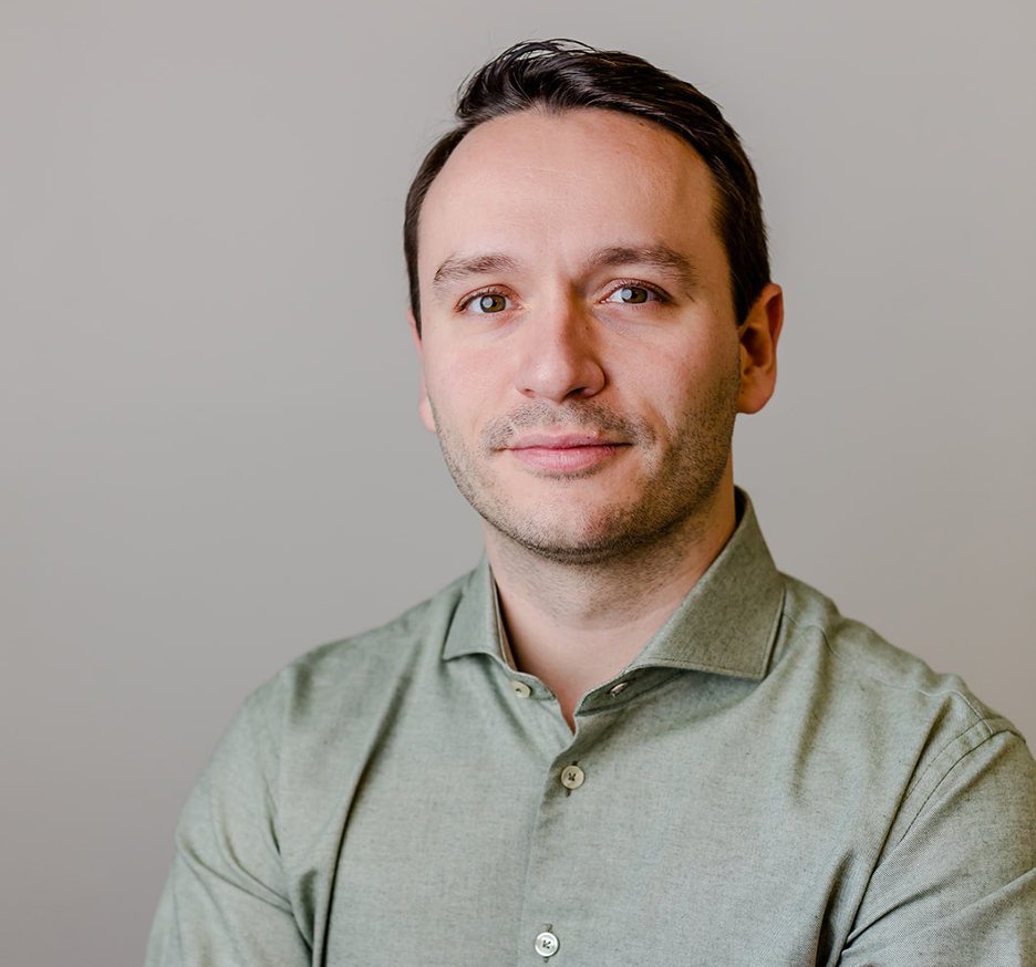 Portrait of a man with brown hair wearing a green button-up shirt against a plain gray background.
