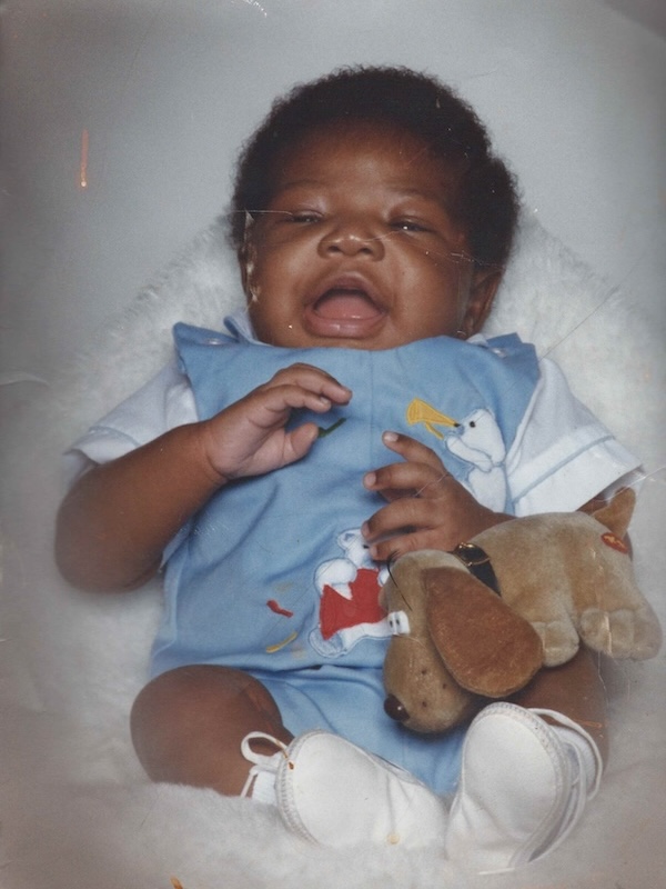 Smiling baby Kelvin wearing a blue romper with a bear design, holding a plush toy dog and wearing white shoes.
