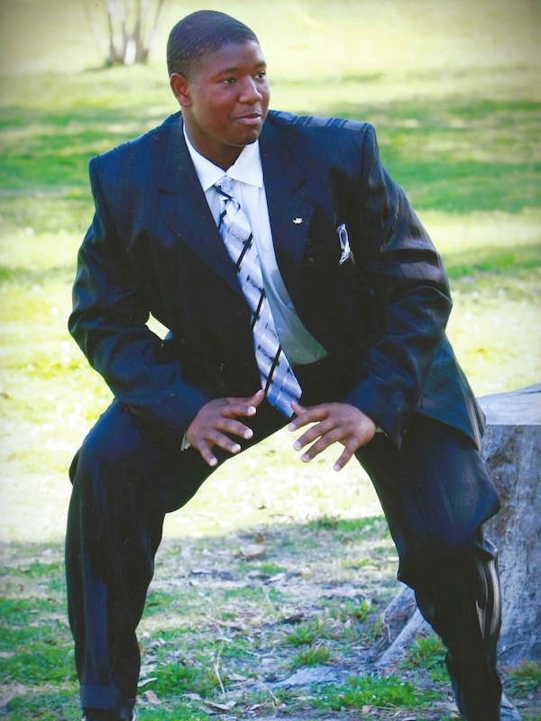 Young Kelvin in a black suit and striped tie crouching on grass outdoors.