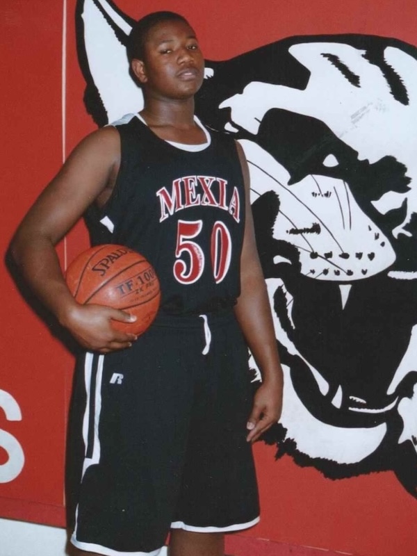 Young Kelvin as a basketball player wearing black Mexia jersey number 50 holding a basketball, standing in front of a large panther mural.