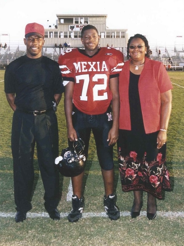 Young Kelvin as a football player in Mexia red jersey number 72 standing on a football field between his dad in black and his mum in a red jacket and floral skirt.