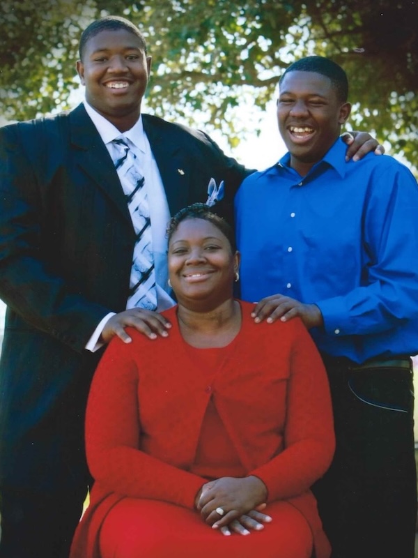 Kelvin's mum in a red dress sitting outdoors with kelvin and his brother standing behind her, all smiling.