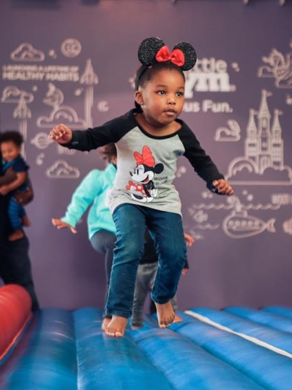 Kalena wearing Minnie Mouse ears headband and shirt jumping on a blue inflatable bouncy castle indoors.