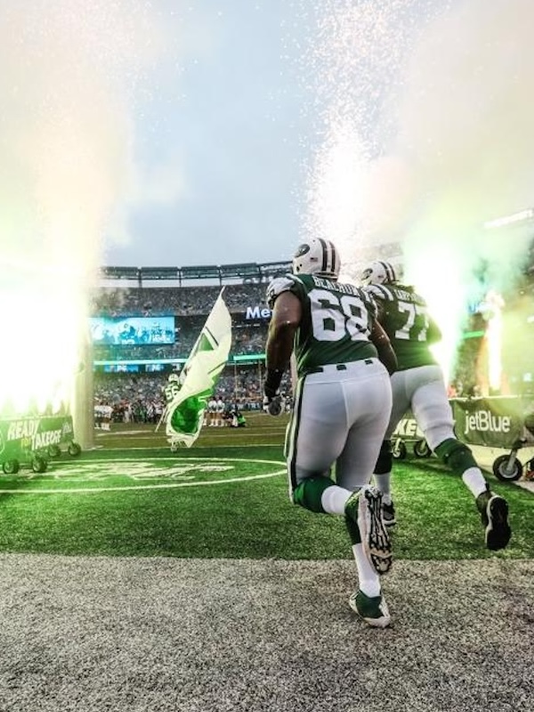 Two football players in green and white uniforms running onto the field amid celebratory smoke and sparks, with a crowd in the stadium.