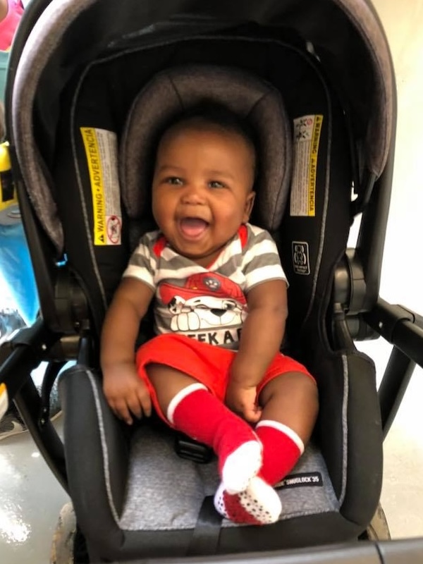 Smiling baby Kelvin III wearing red shorts and matching red socks sitting in a black car seat.