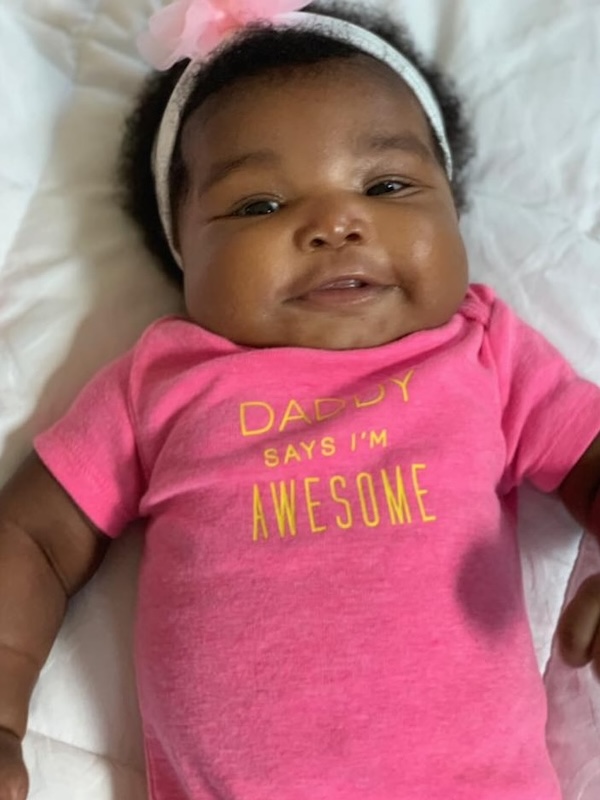 Smiling baby Janelle wearing a pink shirt with yellow text reading 'Daddy says I'm awesome' and a white headband with a pink bow.