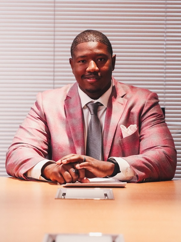 Kelvin in a pink blazer and gray tie sitting at a conference table with hands clasped and blinds in the background.