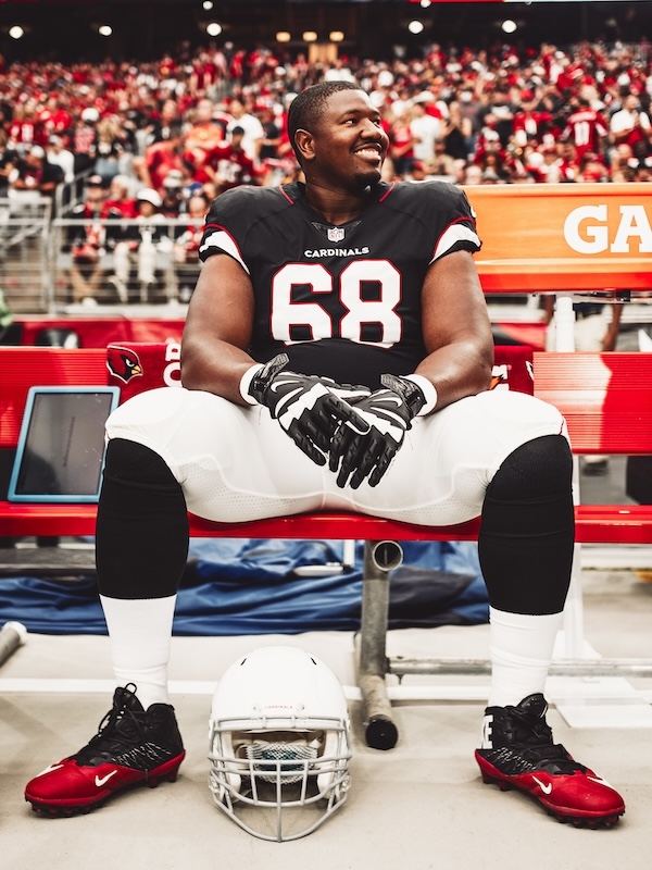 Kelvin wearing number 68 Cardinals uniform sitting on a bench with helmet on the ground in front, smiling and looking to the side.