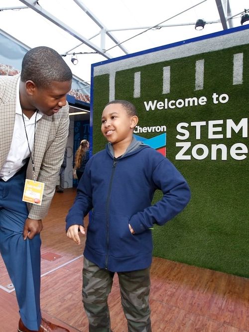 Kelvin in business attire talks to smiling boy in blue jacket and camo pants in front of a STEM zone sign.