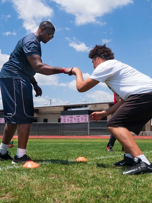 Two young men on a sports field engaging in a handshake or fist bump over grass with orange training cones nearby.