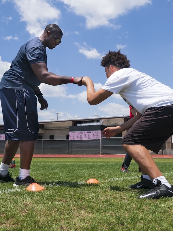 Two young men on a sports field engaging in a handshake or fist bump over grass with orange training cones nearby.